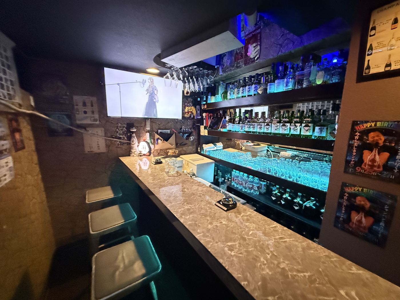 BAR NOMENOMEの店内。青く光るボトル棚とグラス棚の前に大理石調のカウンターと椅子が並び、奥にカラオケの画面